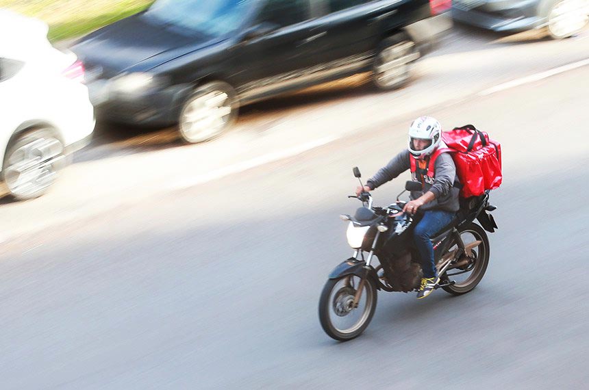 Entregador em motocicleta, no trânsito, com a típica caixa de entregas vermelha nas costas