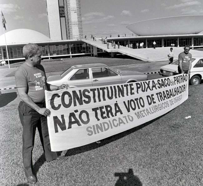 Dois trabalhadores estendem faixa em frente ao Congresso Nacional, durante a Constituinte de 1988. A faixa diz: Constituinte puxa-saco de patrão não terá voto de trabalhador. Sindicato Metalúrgicos de Osasco.