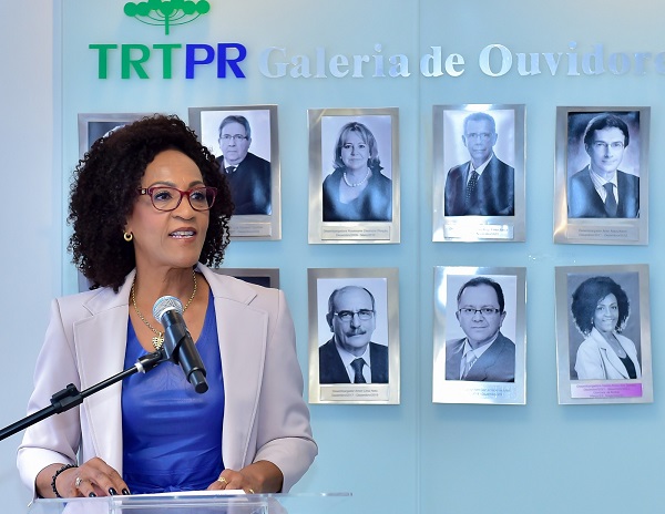 A imagem mostra a desembargadora Neide Alves dos Santos (mulher negra com vestido azul e terno feminino branco) do lado direito. Ela discursa diante das fotos da galeria de ouvidores do TRT-PR. Sua expressão é de alegria.