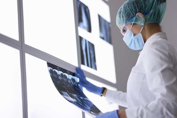 Fotografia de uma mulher em um ambiente hospitalar. Ela usa jaleco branco, touca, máscara e luvas. Nas mãos, ela segura um rádio-x em frente a um monitor iluminado.