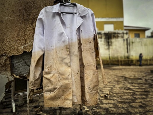 Fotografia de um jaleco branco, utilizado por profissionais de saúde, pendurado com um cabide em uma parede suja. A metade de baixo do jaleco está sujo de lama.