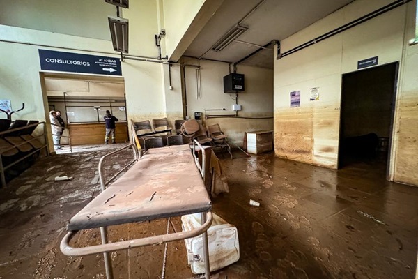 Fotografia mostra o interior do Centro de Saúde Santa Marta, em Porto Alegre. Em primeiro plano, uma maca completamente suja de lama. O chão enlameado tem marcas de botas. As paredes e os móveis estão sujos de lama.