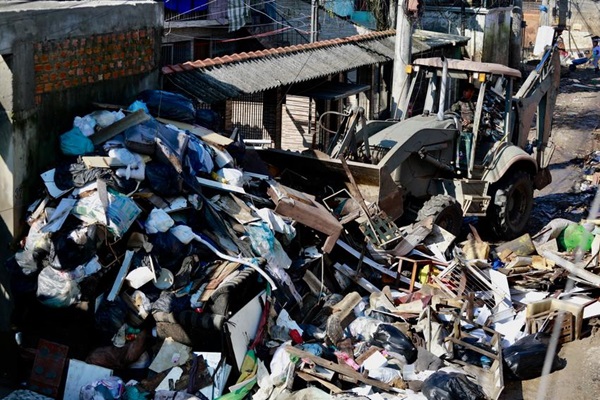 Fotografia do alto mostra uma pilha de entulhos sendo movimentada por uma retroescavadeira do Exército Brasileiro. A máquina fica pequena perto da montanha de lixo. O trabalho ocorre em uma rua com casas simples, sem reboco nas paredes.