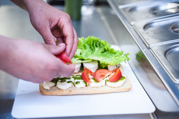 Fotografia das mãos de uma pessoa manipulando pedaços de tomate para colocar dentro de um sanduíche. Pão, que já tem internamente pedaços de queijo, alface e um molho, está sobre uma tábua branca plástica, que está sobre uma bancada de alumínio. 