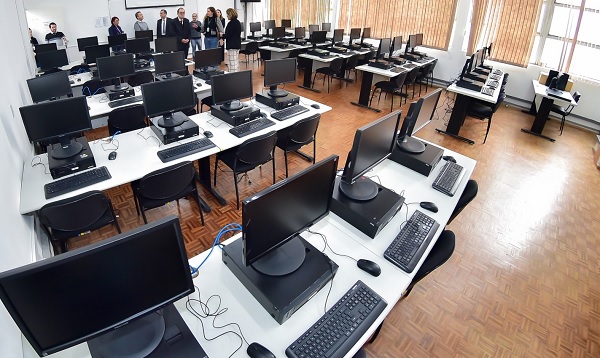 Fotografia da perspectiva do fundo de uma sala de aula com dez bancadas de mesas, cada uma com cinco computadores e periféricos, com cinco bancadas de cada lado, separadas por um corredeor ao centro. Ao fundo, um grupo de pessoas visita a sala. 