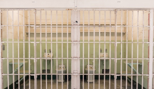 Fotografia de duas celas vazias fechadas com grades de ferro pintadas na cor branca. Em cada cela, um sanitário, uma pia e dois suportes de madeira para serem usados como assento ou cama. 