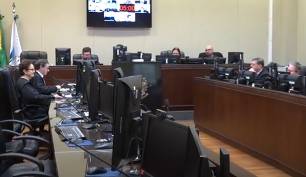 Fotografia da sala de julgamento da 5ª Turma com duas bancadas com computadores frente a frente, ocupada por quatro desembargadores, três homens e uma mulher. E ao fundo a mesa de Presidência da Turma, perpendicular às bancadas, e ocupada por quatro pessoas, três homens e uma mulher.