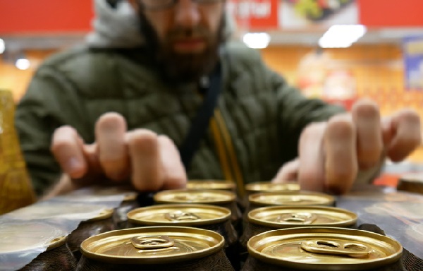 Fotografia da perspectiva de uma prateleira de uma loja. Em primeiro plano, a parte superior de várias latas. Ao fundo, desfocado, um homem com barba leva as mãos para as latas com menção de pegar uma lata em cada mão.