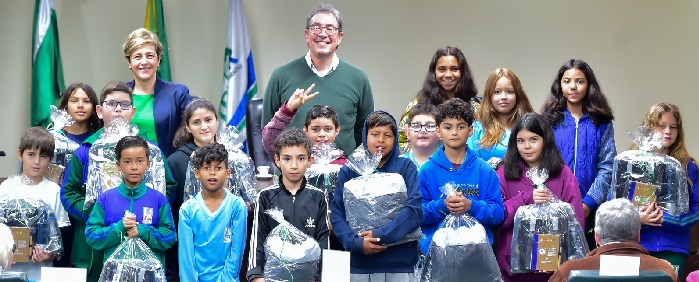 Fotografia mostra 16 crianças juntas segurando os kits do projeto nas mãos. Entre elas, a juíza auxiliar da Presidência, Angélica Cândido Nogara Slomp, mulher branca de cabelos loiros curtos; e o secretário de Família e Desenvolvimento Social de Almirante Tamandaré, Ernesto Antonio Rossi, homem branco de cabelos parcialmente grisalhos e óculos. 