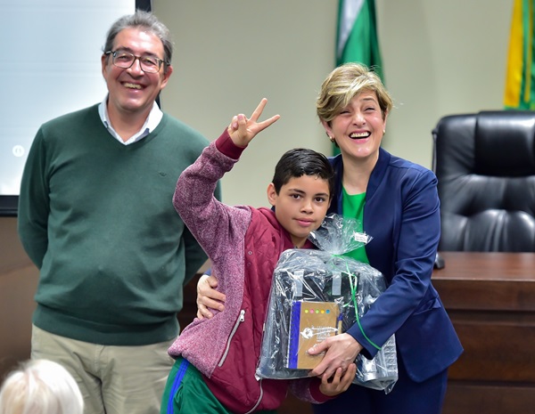 Fotografia mostra um menino de pele branca e cabelos pretos recebendo o kit do projeto com uma mão e com o outro braço erguido faz sinal V com os dedos. Ao lado dele, a juíza auxiliar da Presidência, Angélica Cândido Nogara Slomp, entrega o kit e ri efusivamente. Ela é uma mulher branca de cabelos loiros curtos. No outro lado do menino, está o secretário de Família e Desenvolvimento Social de Almirante Tamandaré, Ernesto Antonio Rossi, homem branco de cabelos parcialmente grisalhos e óculos. Ele ri.