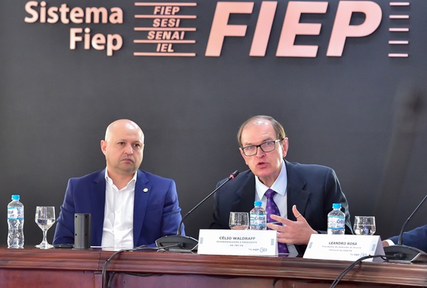 Dois homens sentados em frente a uma mesa de madeira com copos e garrafas plásticas de água sobre ela. Do lado direito, o presidente da Fiep, Edson Vasconcelos, homem branco, calvo, de terno azul e camisa branca. Do lado direito, o presidente do TRT-PR, Célio Waldraff, fala em um microfone. Ele é branc, parcialmente calvo, de óculos, usa terno azul escuro e gravata lilás.