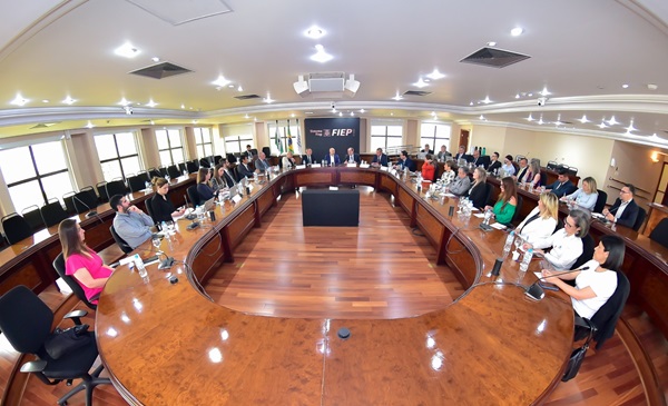 Grupo de aproximadamente 30 pessoas sentadas em torno de uma mesa de madeira em formato oval com painel ao fundo da Fiep.