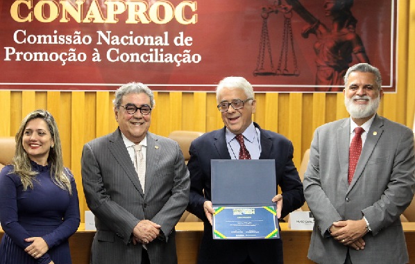 Fotografia de quatro pessoas perfiladas lado a lado, três homens e uma mulher. Ao fundo, cartaz escrito Conaproc, Comissão Nacional de Promoção à Conciliação. Da esquerda para a direita, a juíza Auxiliar da Vice-Presidência do TST Roberta de Melo Carvalho, mulher branca de cabelos loiros compridos; mnistro vice presidente do TST Aloysio Corrêa da Veiga, homem branco de cabelos grisalhos e óculos; desembargador do TRT-PR Eliázer Antonio Medeiros, homem branco grisalho de óculos, ele segura o diploma de premiação; e o ministro presidente do TST Lelio Bentes Côrrea, homem branco grisalho.