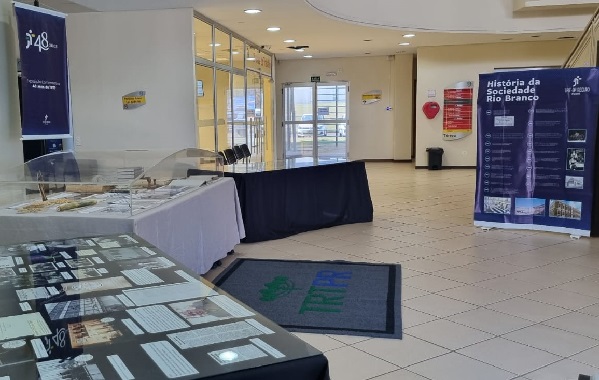 Fotografia do espaço de entrada do Fórum de Maringá. À direita, três bancada de mesas com objetos e documentos expostos. Atrás um banner de 48 anos do TRT-PR. Do lado direito, banner sobre a história da Sociedade Rio Branco, local do prédio sede do Tribunal. 