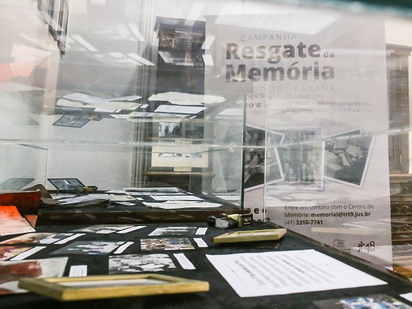 Fotografia de uma mesa com suporte de vidro protegendo objetos em exposição. Ao fundo, cartaz da exposição, com o título, resgate da memória, distorcido pelo vidro. 