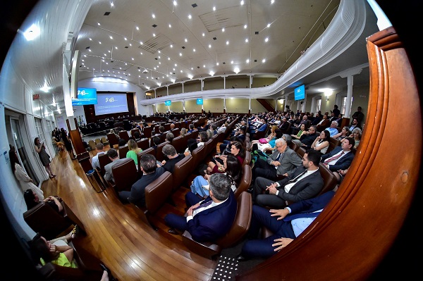 Fotografia do plenário Pedro Ribeiro Tavares em que mostra a plateia parcialmente de costas e ao fundo as mesas com desembargadores e autoridades. Ao fundo, tela azul com símbolo estilizado de 48 anos do Tribunal. Fotog usa lente olho de peixe, que distorce as bordas da imagem.