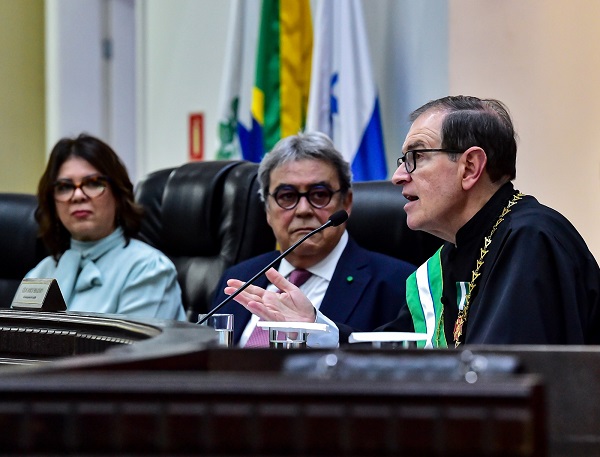 Fotografia do presidente do TRT-PR, Célio Waldraff, discursando em frente a um microfone. Ele usa toga preta, uma faixa, colar e medalha da Ordem do Mérito Judicial das Araucárias. Ao lado dele e ao fundo da foto, assistem o discurso o vice-presidente e presidente eleito do TST, ministro Aloysio Corrêa da Veiga, e asub procuradora-geral para assuntos de planejamento institucional do Ministério Público do Paraná (MP-PR), Terezinha de Jesus de Souza Signorini.