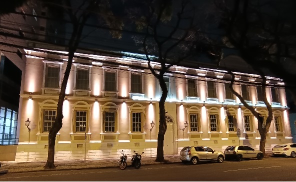 Fachada do Casarão à noite com iluminação na cor amarela em todo o prédio, destancando as duas linhas de janelões. Em frente, árvores e carros estacionados.