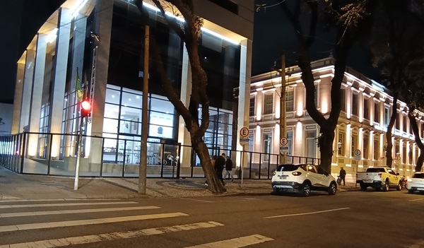 Fachada lateral do prédo sede do TRT-PR com colunas altas de concreto e o prédio com vidro espelhado. Ao lado o casarão histórico, ambos iluminados na cor amarela.