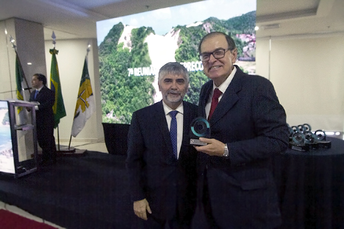 Fotografia do juiz e secretario geral dp CSJT Bráulio Gusmão ao lado do presidente do TRT-PR, desembargador Célio Waldraff. Ambos vestem terno, Braulio de gravata azul e Célio de gravata vermelha. O presidente do TRT-PR segura um trofeu com base em madeira e uma simbolo circular em acrílico.