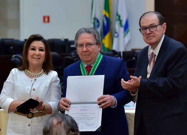 Fotografia de dois homens e uma mulher em pé, uma ao lado da outra, posam para a fotografia. Da esquerda para a direita, a desembargadora Ana Carolina Zaina, veste casaco branco e usa colares; desembargador Luiz Eduardo Gunther, veste terno azul co gravata vermelha, ele tem em mão um diploma e um cola com a homenagem, com fita verde e branca; o desembargador presidente do TRT-PR, Célio Waladraff, de terno azul com gravata vermelha e branca, ele aplaude o momento.