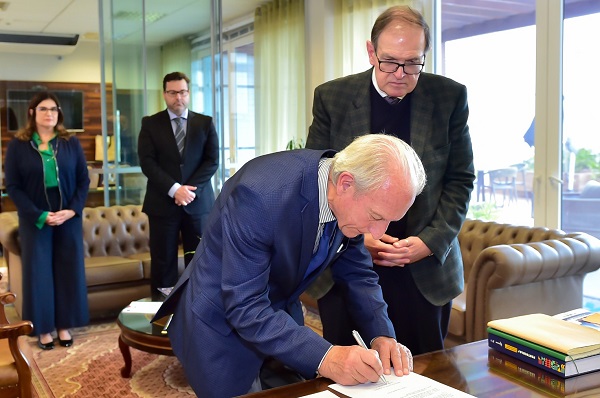 Fotografia mostra assinatura do presidente da ACP, Antonio Deggerone, à campanha. Ele está curvado com caneta na mão assinando o documento sobre mesa de madeira. Ao lado, o presidente Célio Waldraff. Ao fundo, o desembargador eleitoral do TRE-PR José Rodrigo Sade e a vice procuradora-chefe do MPT-PR, Patrícia Blanc Gaidex.