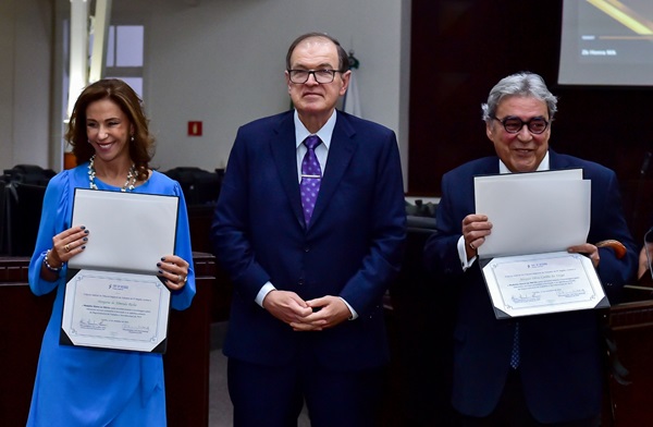 Fotografia da homenagem à ministra Morgana Richa e ao Ministro Aloysio Corrêa. Entre os dois, o presidente do TRT-PR, desembargador Célio Waldraff. A ministra Morgana é mulher branca de calebos compridos até o ombro e veste vestido azul. Ela segura nas mãos um diploma. Desembargador Célio é homem branco, usa óculos, parcialmente calvo, veste terno azul escuro com gravata lilás. O ministro Aloysio é um homem branco de cabelos grisalhos, usa óculos e veste terno azul. Ele segura um diploma nas mãos.