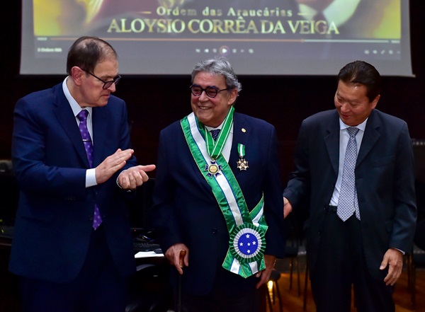 Fotografia do momento da entrega da Comenda das Araucárias ao ministro Aloysio. Do lado esquerdo da image, o presidente do TRT-PR, desembargador Célio Waldraff. No centro, o ministro Aloysio com a medalha e a faixa da Comenda das Araucárias. A faixa está sobre o tronco do ministro, é da cor verde com uma faixa branca no meio e a junção tem o símbolo do Cruzeiro do Sul. No lado direito da imagem, o ex-ministro Fernando Eizo Ono. Todos vestem ternos.