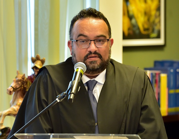 Fotografia frontal do juiz José Alexandre Barra Valente, homem pardo de barba grisalha, cabelos curtos e quye usa óculos. Ele veste terno com gravata azul e sobre a roupa a toga de magistrado. Ele fala em frente a um microfone e uma tribuna na solenidade de posse.