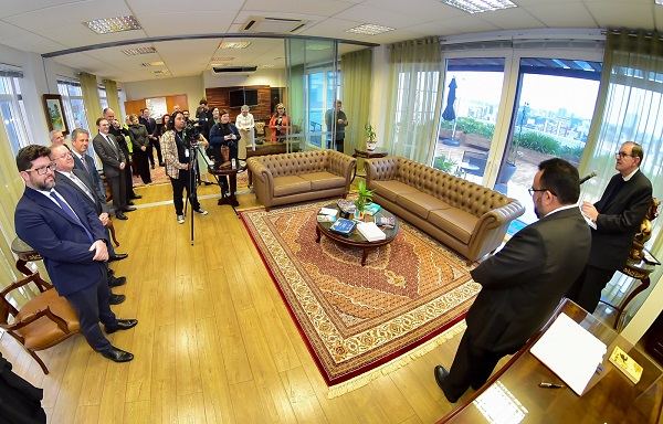 Fotografia com leite olho de peixe (que distorce levemente as extremidades para auimentar o ângulo) do Gabinete da Presidência. À direita, o presidente do TRT-PR, desembargador Célio Waldraff, e o magistrado José Alexandre Barra Valente. À esquerda, grupo de pessoas, entre desembargadores, servidores do TRT-PR e familaires do magistrado, acompanham a cerimônia.