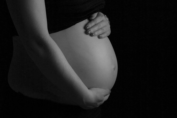 Fotografia sem cor de uma mulher grávida com as duas mãos da barriga, a direita embaixo e a esquerda na parte superior. A imagem mostra a mulher de lado com enquadramento do tronco, entre o ombro e a cintura.