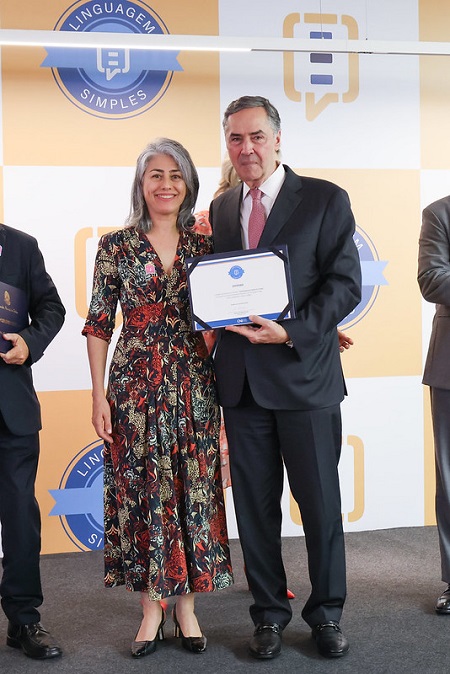 Fotografia frontal da juiza auxiliar da Presidência do TRT-PR, Simone Galand de Figueiredo, mulher branca de cabrelos grisalhos e vestido florido, recebe o selo da linguagem simples do presidente do STF e do SNJ, ministro Luiis Roberto Barroso, homem branco de cabelos parcialmente grisalhos, que veste tenro ciunza escuro com camisa branca e gravata rosa.