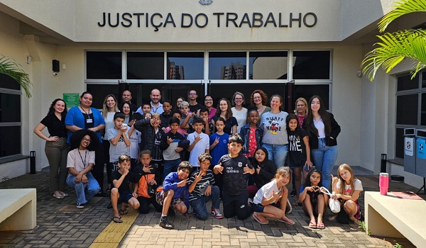 Fotografia frontal de um grupo de crianças com servidoras(es) e magistradas da Justiça do Trabalho em Foz do Iguaçu em frente ao Fórum Trabalhista da cidade.