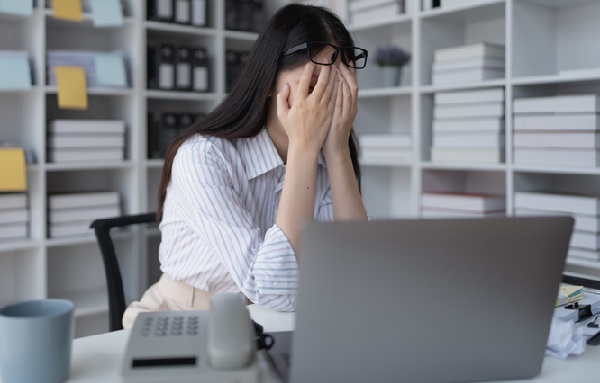 Mulher sentada em cadeira em frente a um laptop cobre o rosto com as duas mãos em um ambiente de escritório.