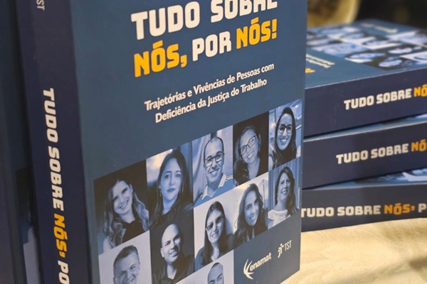 Livro Tudo sobre nós, por nós. Há um livro em pé e outros empilhados, sobre uma mesa. Capa azul escuro com fotos das pessoas que prestaram depoimentos à obra. 