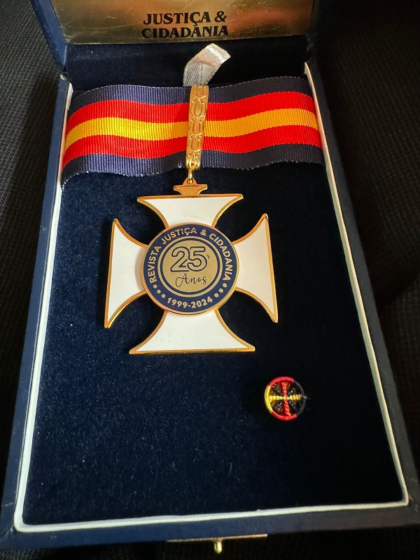 Fotografia da medalha em uma caixa aveludada. Ela tem cordão azul escuro com duas faixas vermelhas e uma amarela ao centro. A medalha tem quatro pontas e ao centro um círculo com os dizeres: Resvita Justiça e Cidadania, 1999-2024, 25 anos.