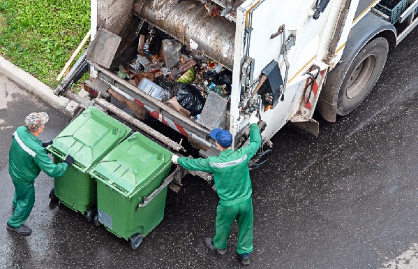 Fotografia do alto mostra dois trabalhadores com uniformes verdes carregando lixo doméstico misto em caminhão de coleta de lixo.