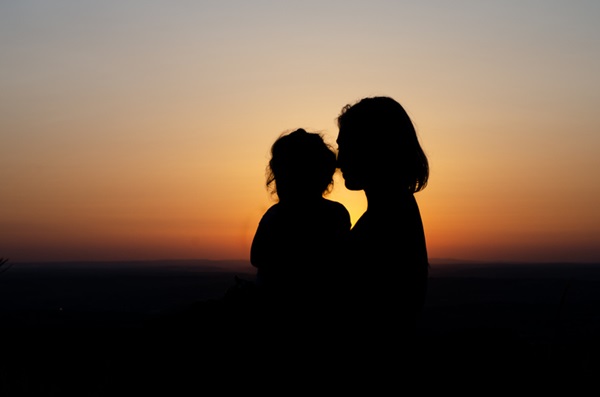 Mãe e filha juntas em um pôr do sol. Iluminação do sol faz perceber apenas o contorno das pessoas.