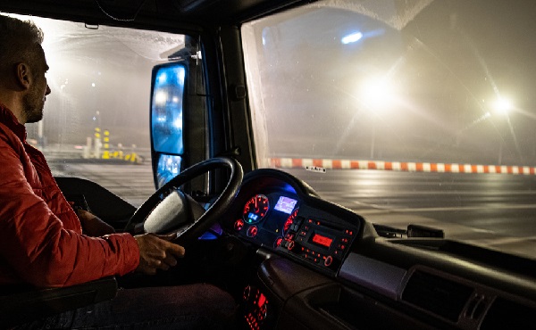 Fotografia de um homem branco na função de motorista ao volante do caminhão durante à noite. Ele está do lado esquerdo da imagem olhando para fora. Ao centro da foto, o volante e painel do veículo E a direita a imagem externa através do para-brisa.