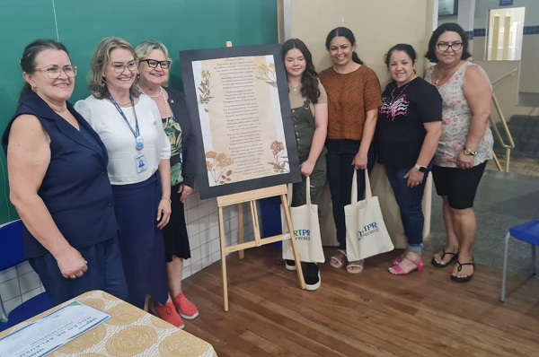 Grupo de sete pessoas perfiladas lado a lado, com um banner sobre um suporte de madeira com um cartaz contendo o texto de uma poesia fixado.