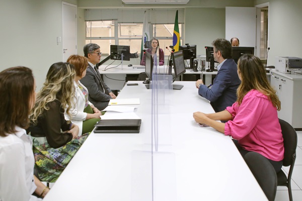 Fotografia de uma sala de audiência com oito pessoas ao todo. No centro, uma mesa com as partes uma de cada lado da mesa, quatro pessoas do lado esquerdo da imagem e duas do lado direito. Ao fundo, outra mesa com a magistrada e um servidor da Justiça de frente. 