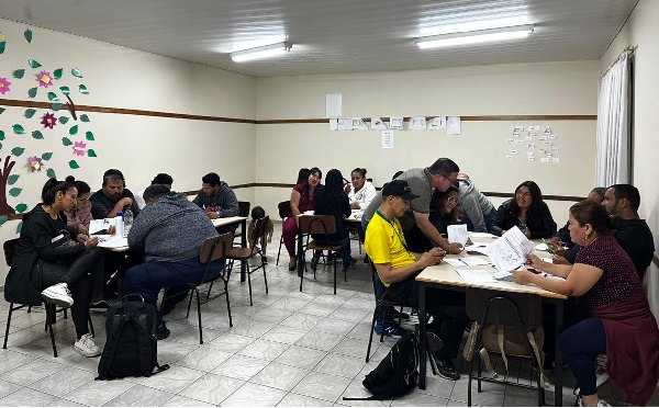 Fotografia de uma sala de aula com três grupos de pessoas sentados em cadeiras em frente a mesa escolares. Cada grupço tem cerca de sete pessoas. Ao fundo, uma parede tem uma colagem em formato de árvore.