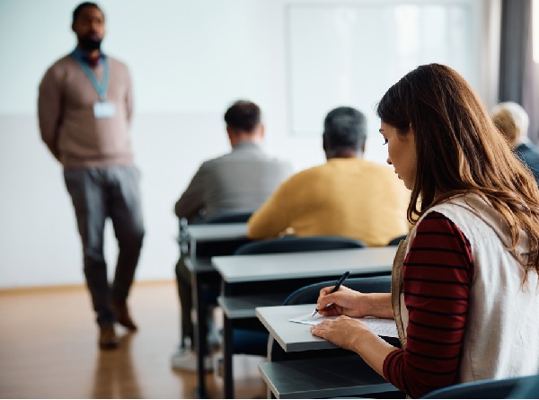 Fotografia de uma sala de aula com uma mulher escrevendo sentada em uma cadeira em frente a uma mesa escolar. Ao fundo, mais linhas de mesas escolares e um homem em pé na posição de professor.