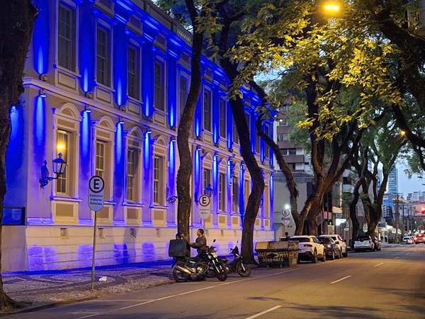 Fotografia da fachada do prédio histórico do TRT-PR, que sediou o Clube Rio Branco, na rua Visconde do Rio Branco com iluminação especial na cor azul. Em frente ao prédio, no entardecer, visão parcial da rua com carros estacionados e um motociclista ao lado de uma moto olhando a fachada.