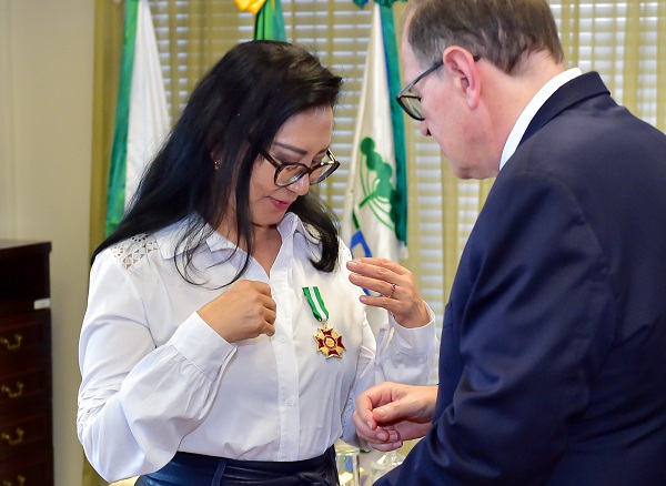 Fotografia da desembargadora Lidia Maejima recebendo a medalha da Ordem das Araucárias do presidente do TRT-PR, desembargador Célio Waldraff. Ela é uma mulher de traços orientais, usa óculos e camisa branca. Medalha está afixada na altura do peito. Em frente dela, o desembagador Célio usa terno cinza escuro.