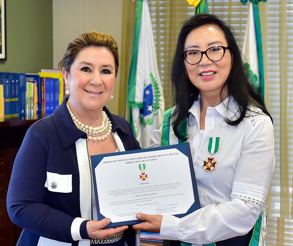 Fotografia da desembargadora Lidia Maejima recebendo o certificado da medalha da Ordem das Araucárias da desembargadora e ex-presidente do TRT-PR, desembargadora Ana Carolina Zaina. Ambas olham para o fotógrafo e seguram o certificado. Maejima é uma mulher de traços orientais, usa óculos e camisa branca. Medalha está afixada na altura do peito. Desembargadora Ana Carolina Zaina é mulher branca, usa blusa azul e colar e brincos de pérolas.