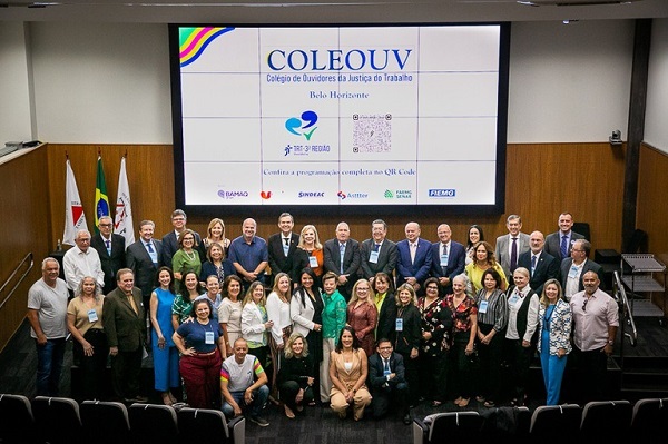 Fotografia do alto de um auditório de um grupo de cerca de 40 pessoas perfiladas lado a lado posando para a foto. Ao fundo, um painel tem escrito Coleouv, colégio de ouvidores do trabalho.