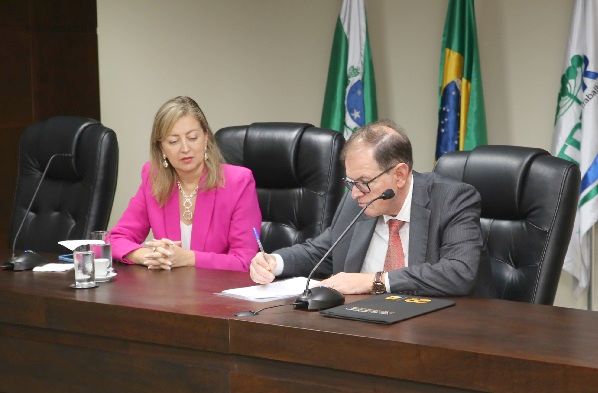 Fotografia do presidente do TRT-PR, desembargador Célio Waldraff, assinando documento sentado em cadeira em frente à mesa de madeira. Ele é um homem branco e veste terno. Ao lado dele, a juíza Sandra Cembranelli Correa veste paleto rosa e tem as mãos cruzadas sobre a mesa ao olhar o momento da assinatura.