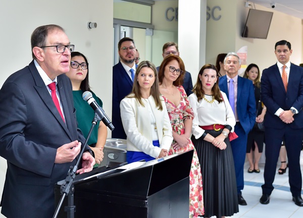 Fotografia lateral em primeiro plano do presidente do TRT-PR, desembargador Célio Waldraff, homem branco com óculos e terno, fala em frente a um microfone e a uma tribuna. Ao fundo, um grupo de pessoas com mulheres e homens assiste o discurso.