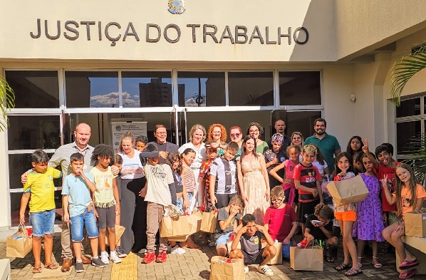 Fotografia de um grupo de cerca de 40 pessoas, a maioria crianças, em frente a entrada do Fórum Trabalhista de Foz do Iguaçu.